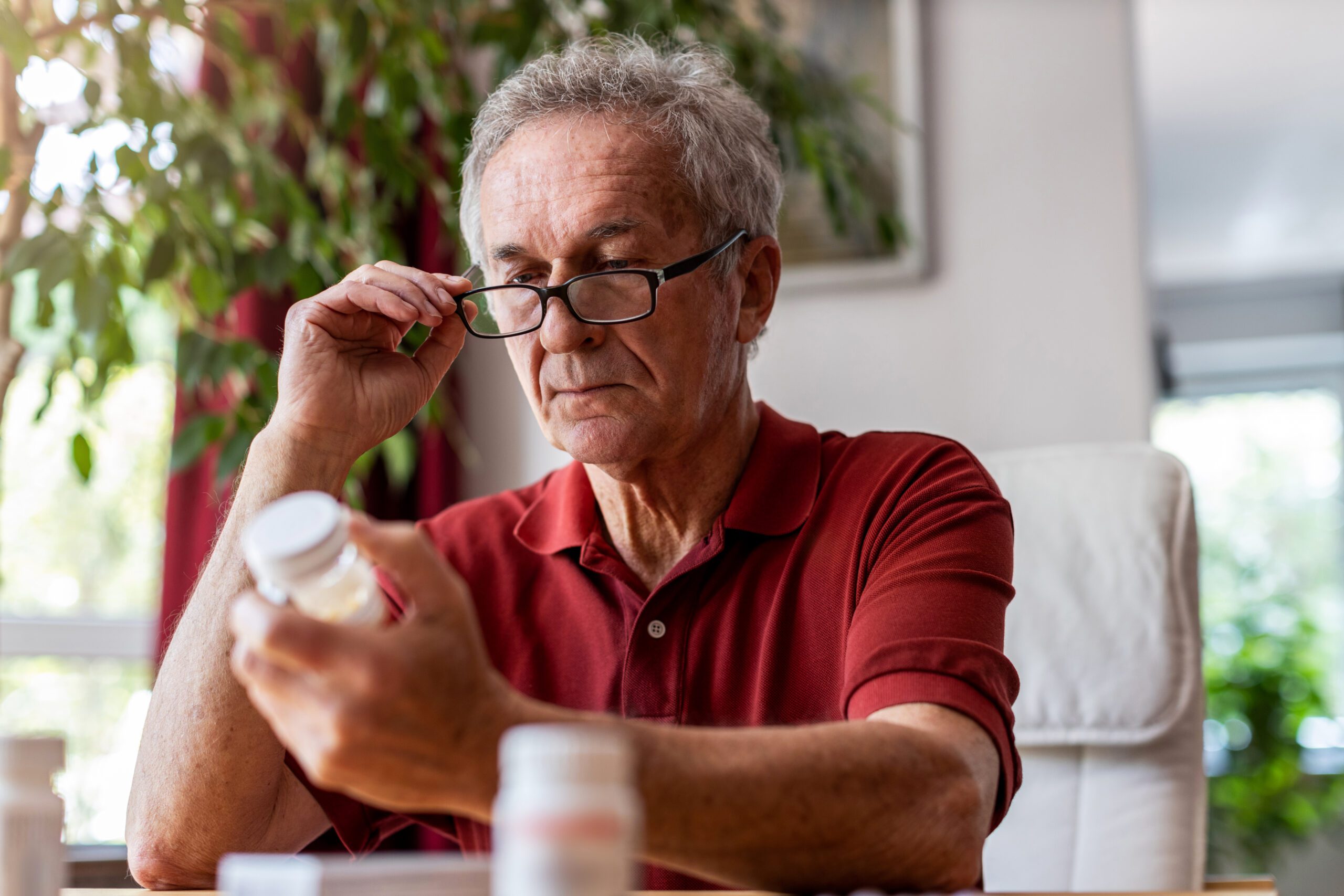 Elderly man checking multiple medication bottles on a table, concentrating and adjusting his glasses